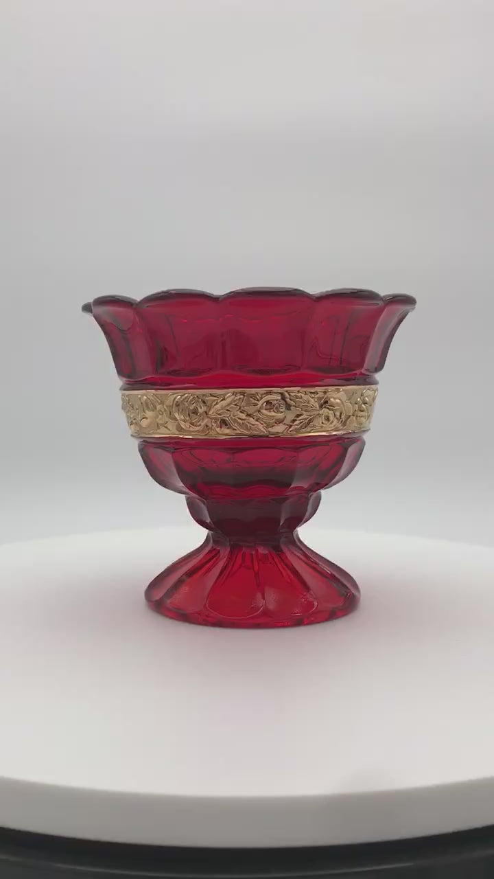 A vintage red glass pedestal bowl with a scalloped rim and a decorative gold band around the middle, set against a plain white background: 1929-1931 Red Ariadne Sugar Basin by Brockwitz. on  rotating display.